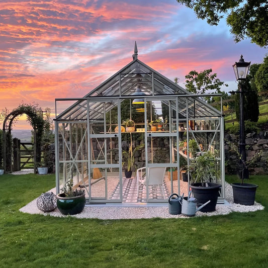 A glass Rhino Greenhouse with potted plants and a chair is situated on a grassy lawn. The background showcases a vibrant sunset and there is a vintage-style lamp post nearby.