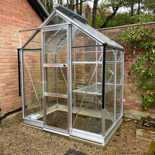 A small glass Rhino Greenhouse stands on gravel, attached to a brick wall, surrounded by greenery. It has a metal frame and a partially open door.