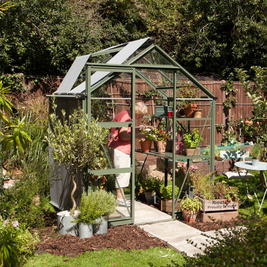 A person tends to plants inside a green metal and glass Rhino Greenhouse with open roof panels. The garden is lush with potted plants, and Rhinogreenhouses boxes are visible nearby.