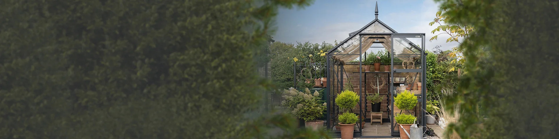 A Rhino Greenhouse stands in a garden, filled with various potted plants and surrounded by lush greenery. The structure features a peaked roof and glass panels, providing a serene gardening space.