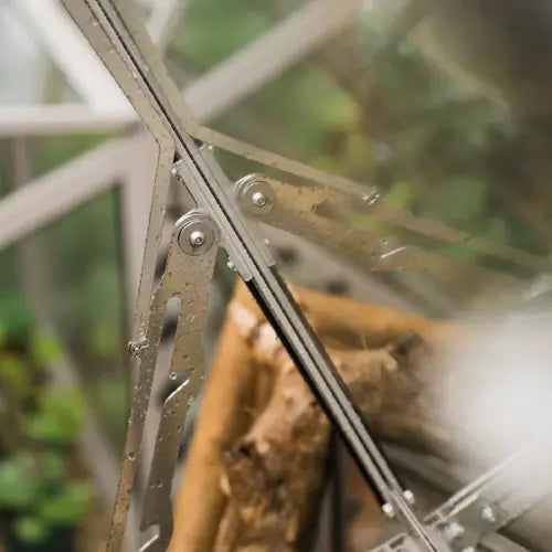 Metal hinge, slightly wet, connects glass panels of a Rhino Greenhouse, surrounded by blurred greenery and wooden elements.