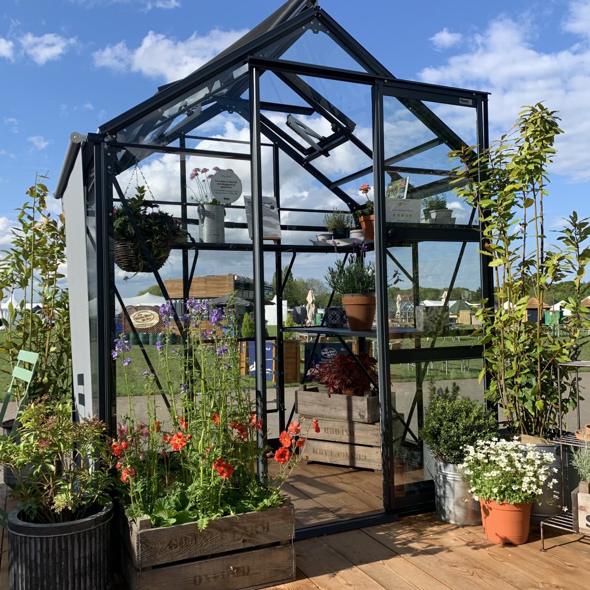 A glass Rhino Greenhouse displays various potted plants and flowers in a garden setting. Sunlight illuminates the interior, and wooden planters and metal containers complement the outdoor greenery and blue sky.