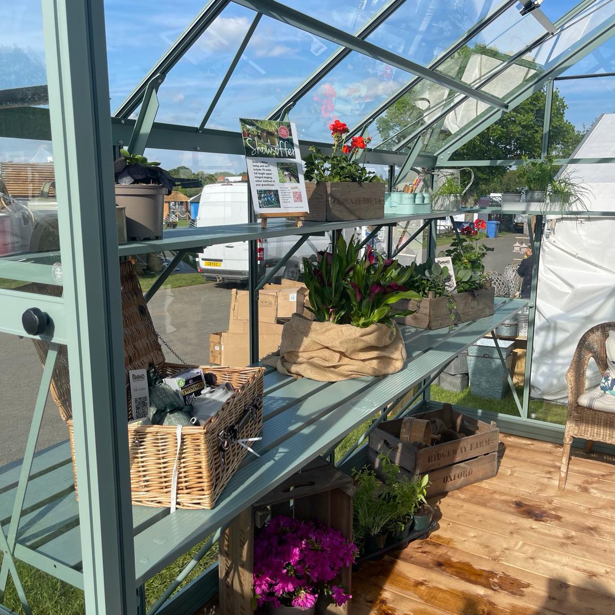 Rhino Greenhouse shelves hold potted plants and gardening accessories. Sunlight filters through glass roof and walls. Nearby, a wicker basket contains items. Background shows outdoor market with vehicles and tents.