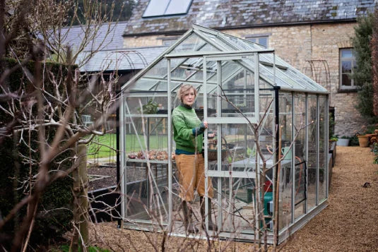 A person stands inside a glass Rhino Greenhouse, holding the door. The setting is a well-maintained garden path surrounded by shrubs, with a stone building in the background.