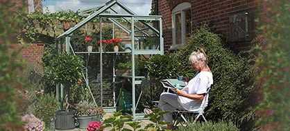 A glass Rhino Greenhouse filled with plants stands in a garden. A person sits nearby on a chair, reading a book. Red brick wall and lush greenery surround the scene.