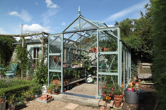 A glass Rhino Greenhouse stands open with plants and gardening tools inside. Surrounding it are flower pots, a small garden, and trees under a clear blue sky.