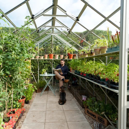 A person sits at a small table reading inside a Rhino Greenhouse filled with various potted plants, surrounded by lush greenery. A cat sits nearby, and a wooden crate labeled Rhino is on the ground.