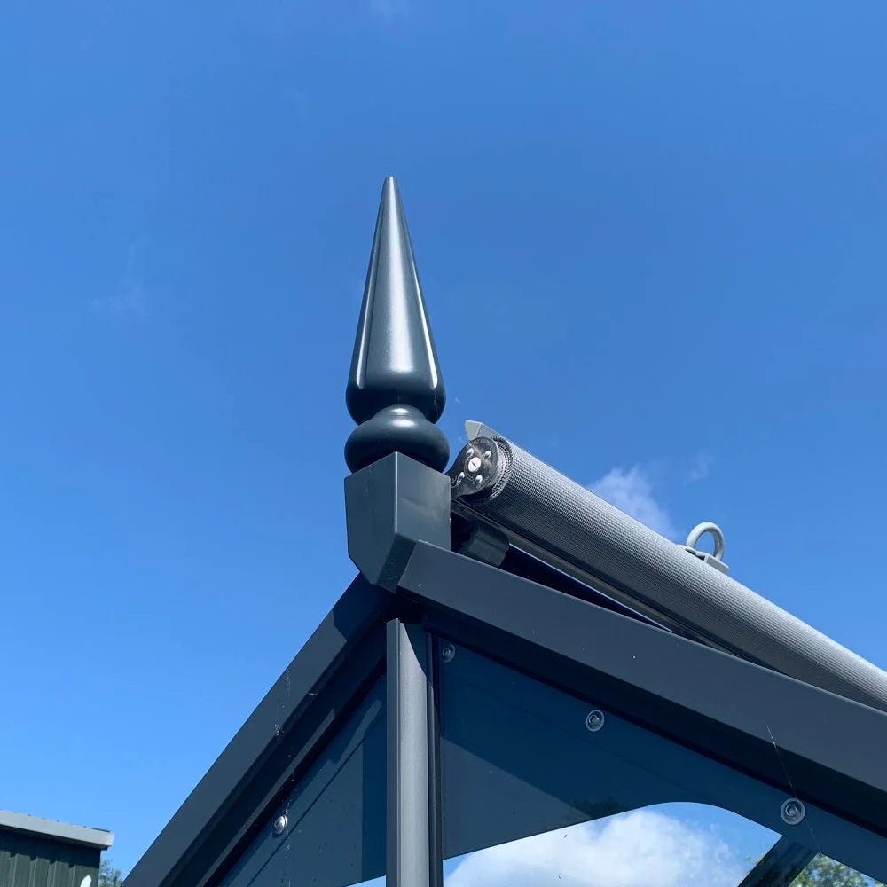 A black metal finial tops a Rhino Greenhouse frame corner, with a rolled-up sunshade attached. The bright blue sky provides a clear, sunny backdrop.