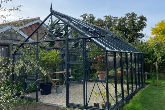 A large glass and metal Rhino Greenhouse houses various potted plants and a small wooden table set, situated in a grassy garden area, with trees and a brick building nearby.