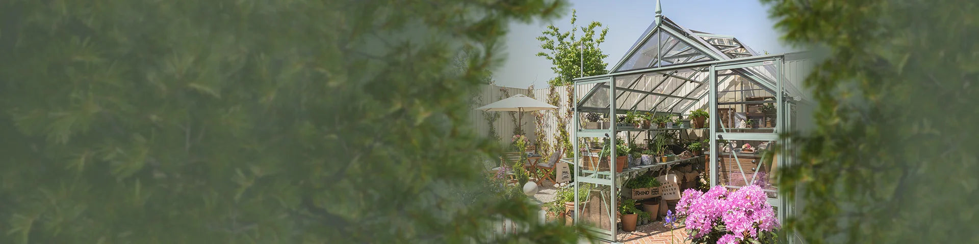 Rhino Greenhouse displays plants and gardening supplies, surrounded by lush garden with trees and flowers; a patio area with chairs and an umbrella is visible nearby, enclosed by a fence.
