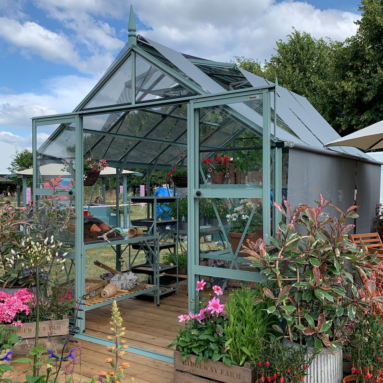 A Rhino Greenhouse with transparent panels and a peaked roof displays potted plants and gardening tools inside. Surrounded by colorful flowers and greenery on a wooden deck, under a partly cloudy sky.