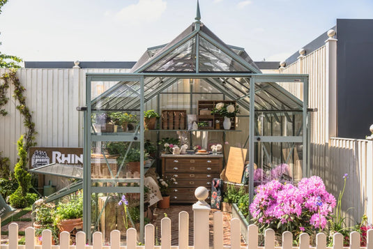 A Rhino Greenhouse with clear glass and metal frames houses plants and gardening tools. It stands on a brick patio, surrounded by a white fence and colorful flowers. Sign reads Rhino Rhino Greenhouses.