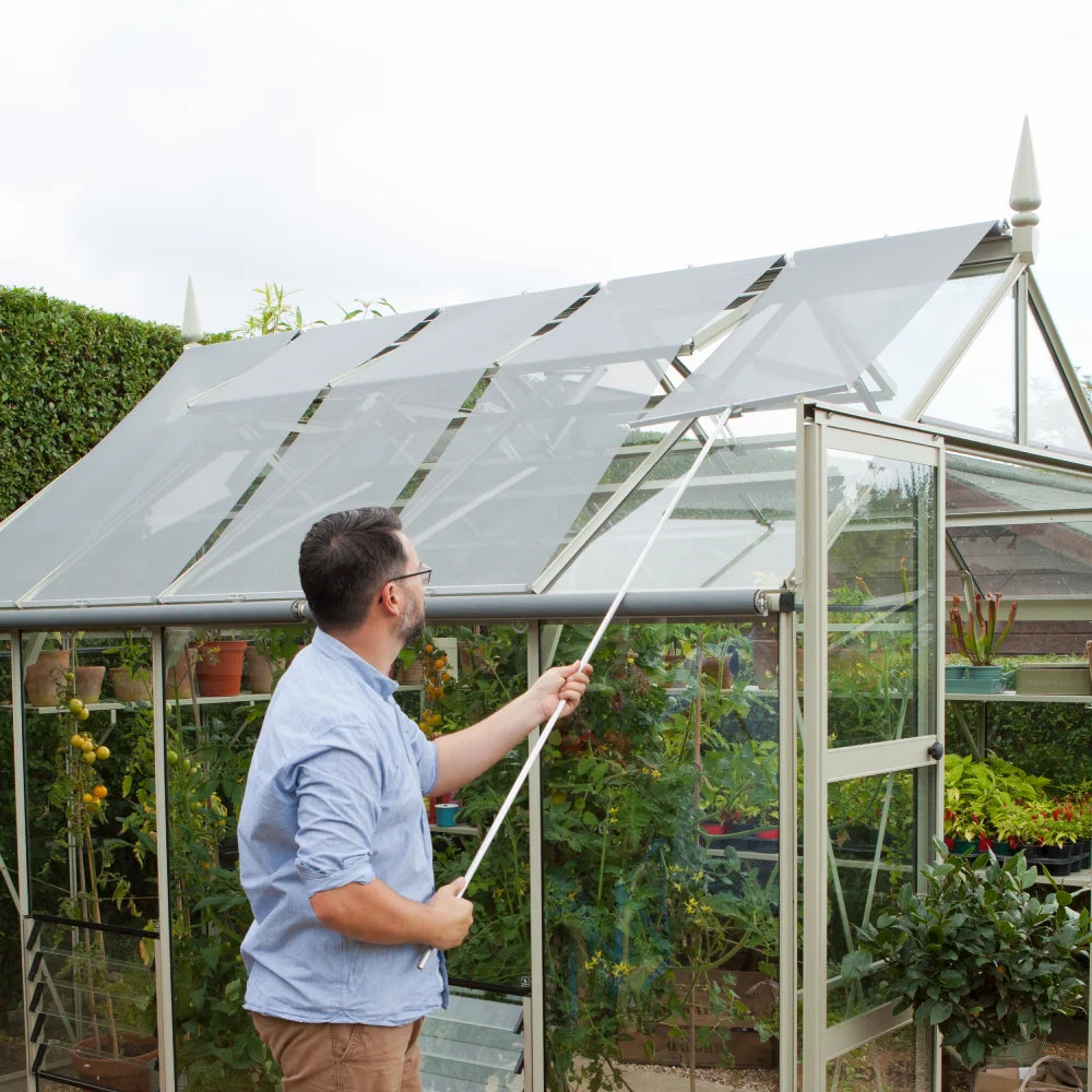 A man adjusts the roof shading of a modern Rhino Greenhouse using a pole, surrounded by lush plants and gardening tools inside, with a hedge in the background.