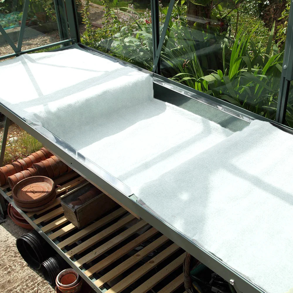 A Rhino Greenhouse bench with white fabric covering. Below, shelves hold stacked terracotta pots and gardening equipment. Sunlight filters through glass panels, illuminating surrounding plants outside.