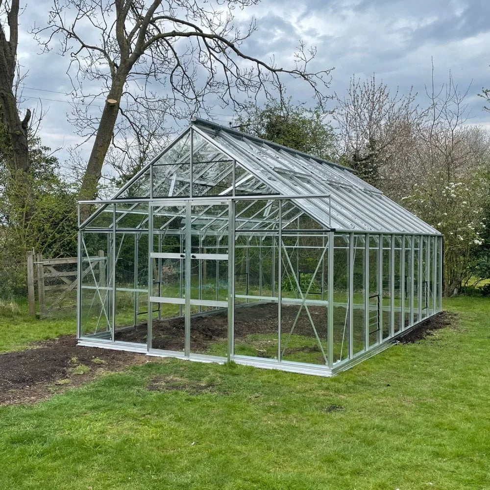 Rhino Greenhouse stands on a grassy lawn, surrounded by trees and shrubs. Its glass panels and metal frame reflect the overcast sky.