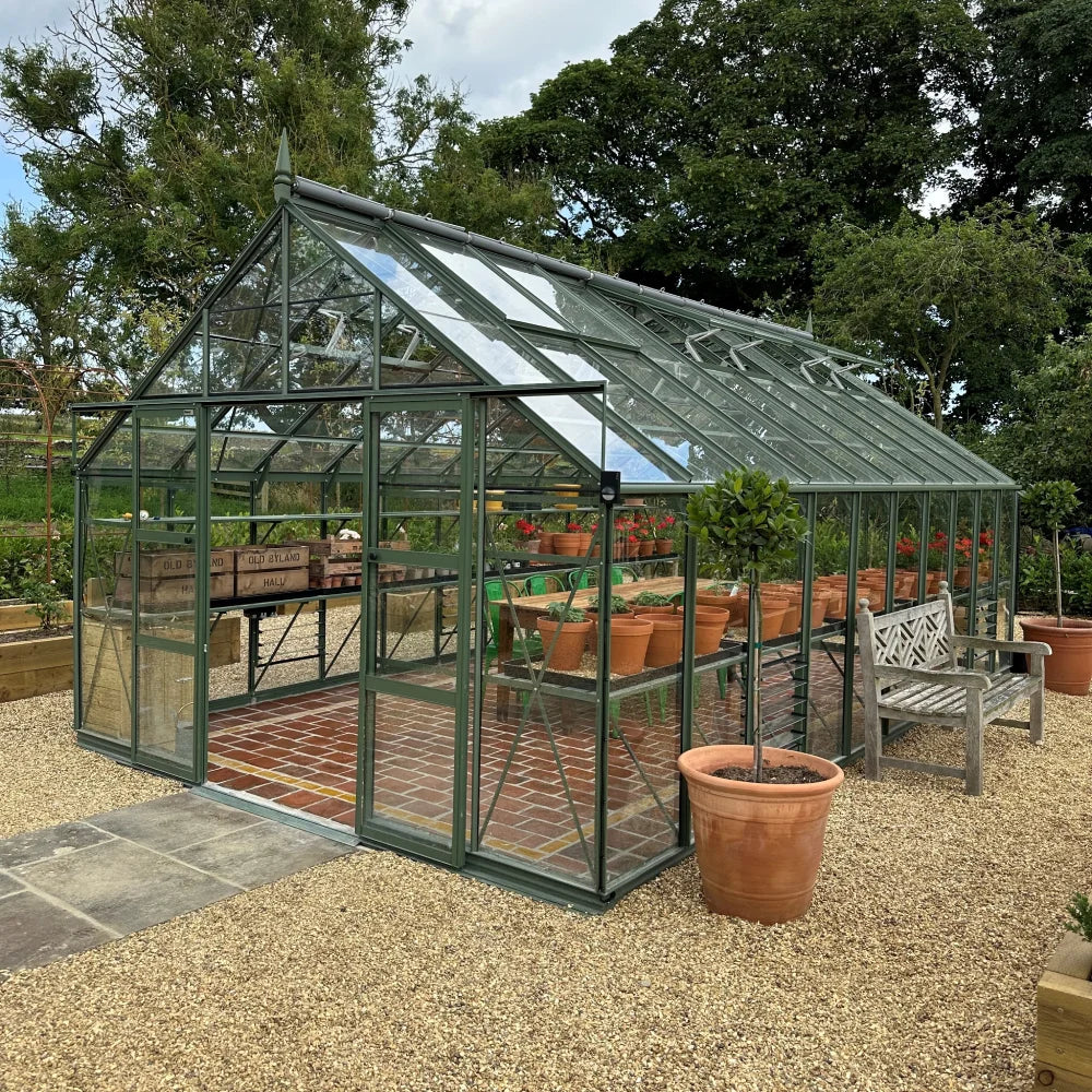 A glass Rhino Greenhouse with a green frame houses rows of potted plants on shelves, surrounded by gravel paths and trees, with a bench and large planters nearby.