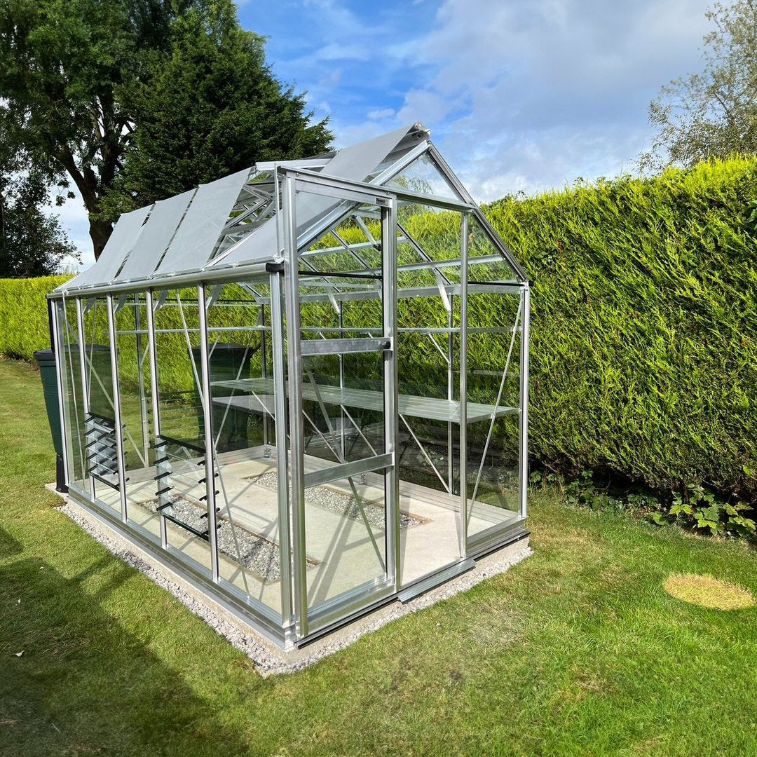 A glass Rhino Greenhouse stands with open roof vents, nestled in a neatly manicured garden, surrounded by lush green hedges and a clear blue sky above.