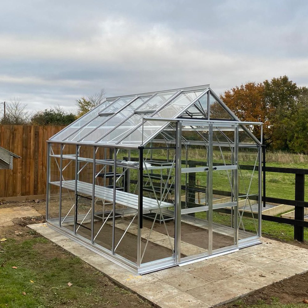 A glass and metal Rhino Greenhouse stands on a paved surface, surrounded by grass and a wooden fence. The sky is overcast, hinting at a typical garden setting.