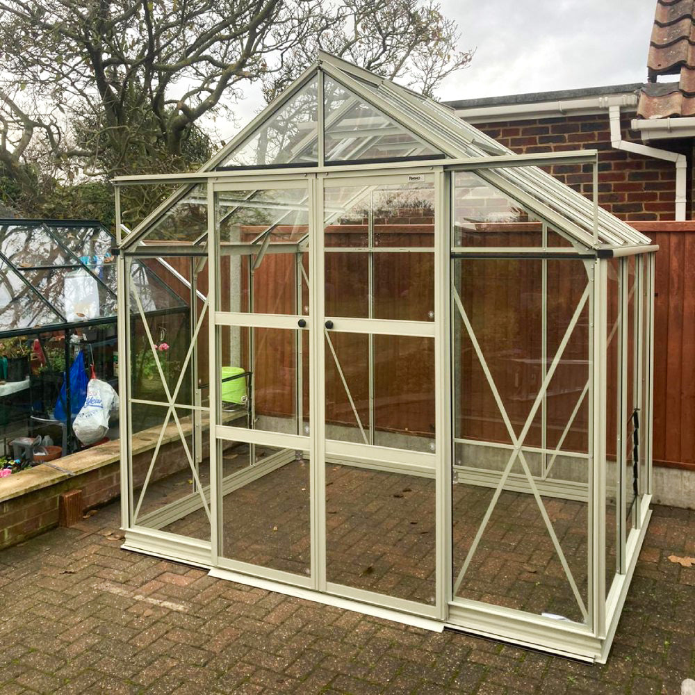 A glass Rhino Greenhouse stands on a brick patio, surrounded by a wooden fence and trees. It has a pitched roof and a visible label reading Rhino above the door.