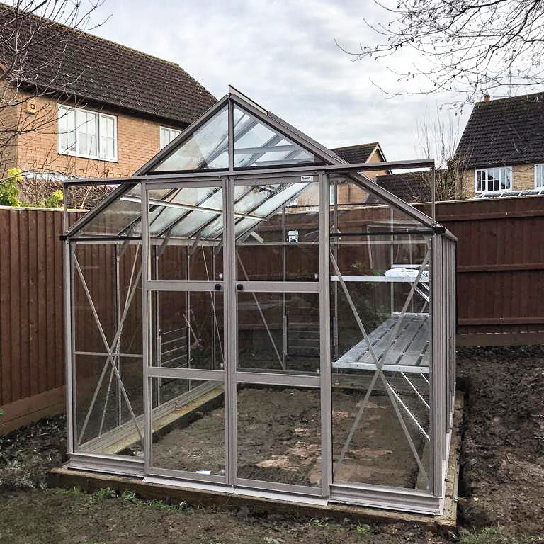 A glass-paneled Rhino Greenhouse stands in a fenced backyard. It features a triangular roof and metal frame, with a soil floor. Nearby, brick houses are visible under overcast skies.