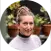 A person with a neat bun hairstyle smiles in front of potted plants, suggesting a gardening setting. The background features blurred foliage and terracotta pots, creating a vibrant, outdoor atmosphere.