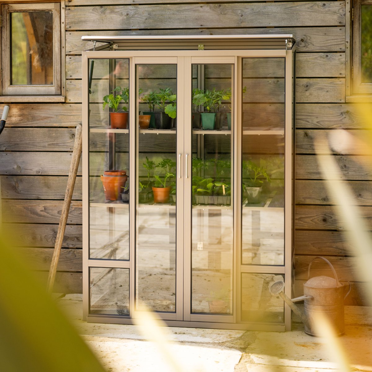 A compact Rhino Greenhouse stands against a wooden wall, containing potted plants on shelves. Nearby, a watering can and a garden tool are visible, suggesting a gardening environment.