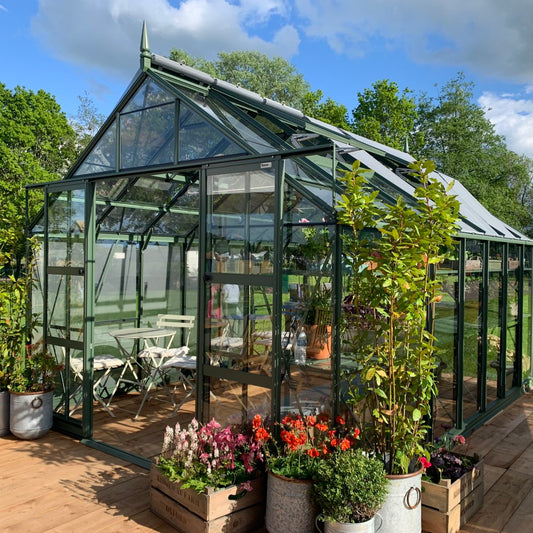 A glass Rhino Greenhouse stands on a wooden deck, surrounded by vibrant potted plants and outdoor furniture. The structure is set in a lush garden under a partly cloudy blue sky.