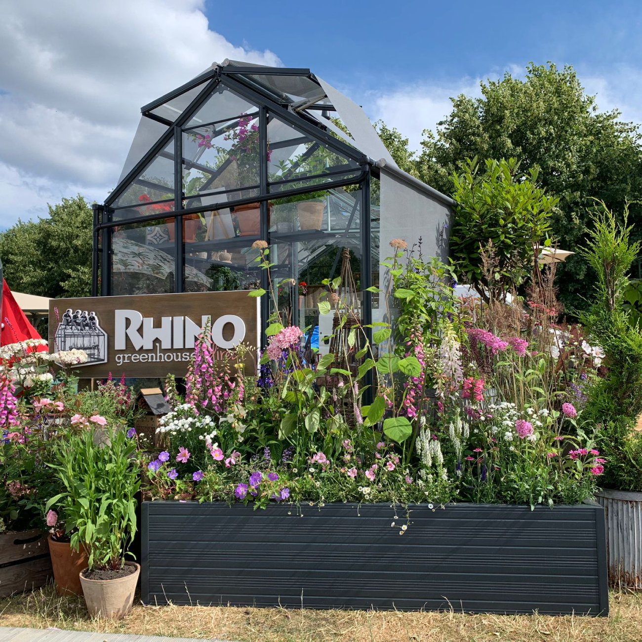 A Rhino Greenhouse displays vibrant plants inside, surrounded by colorful flowers and greenery outside. A sign reads Rhino Rhino Greenhouses against a backdrop of trees and a partly cloudy sky.