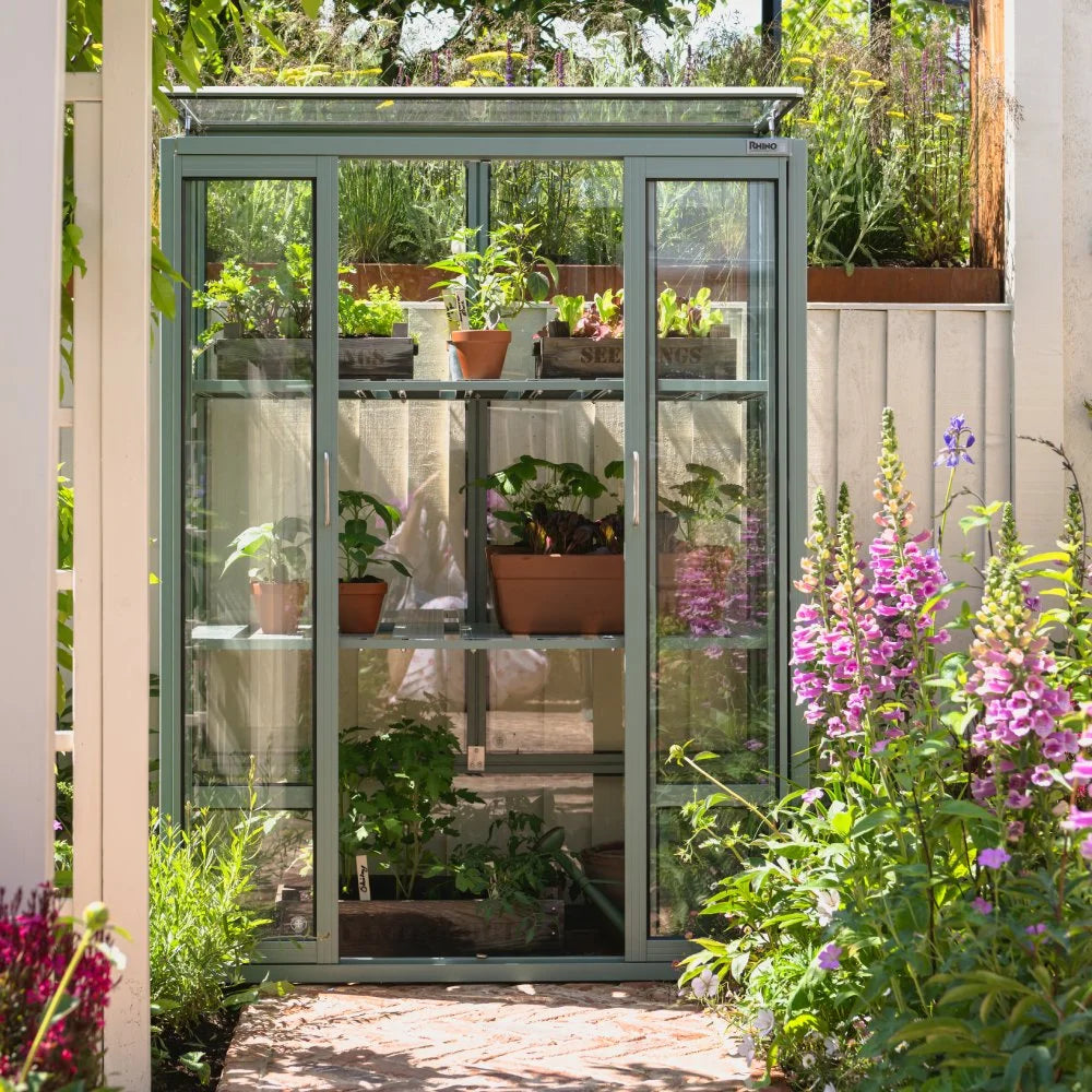 Rhino Greenhouse with glass walls, housing potted plants and small seedlings on shelves. Set amidst a lush garden with colorful flowers, sunlight streaming through the area. Text: Rhino.