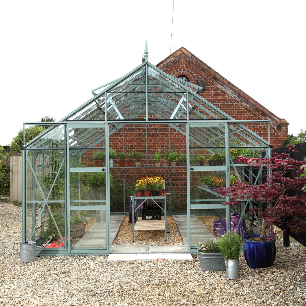 A glass Rhino Greenhouse stands with potted plants inside and outside, located on a gravel path, adjacent to a red brick building and surrounded by lush greenery.