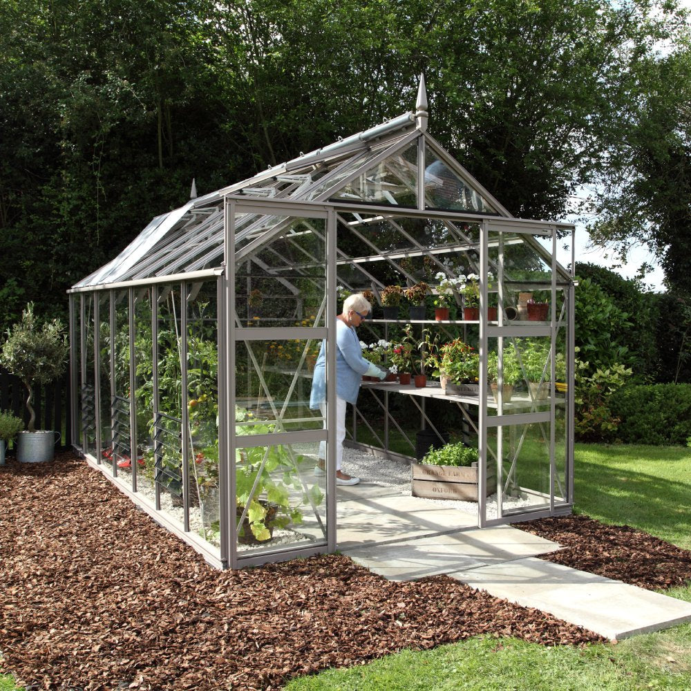 A glass Rhino Greenhouse contains numerous potted plants on shelves. A person tends to them inside, surrounded by lush greenery and a bark-covered path leads to the entrance.