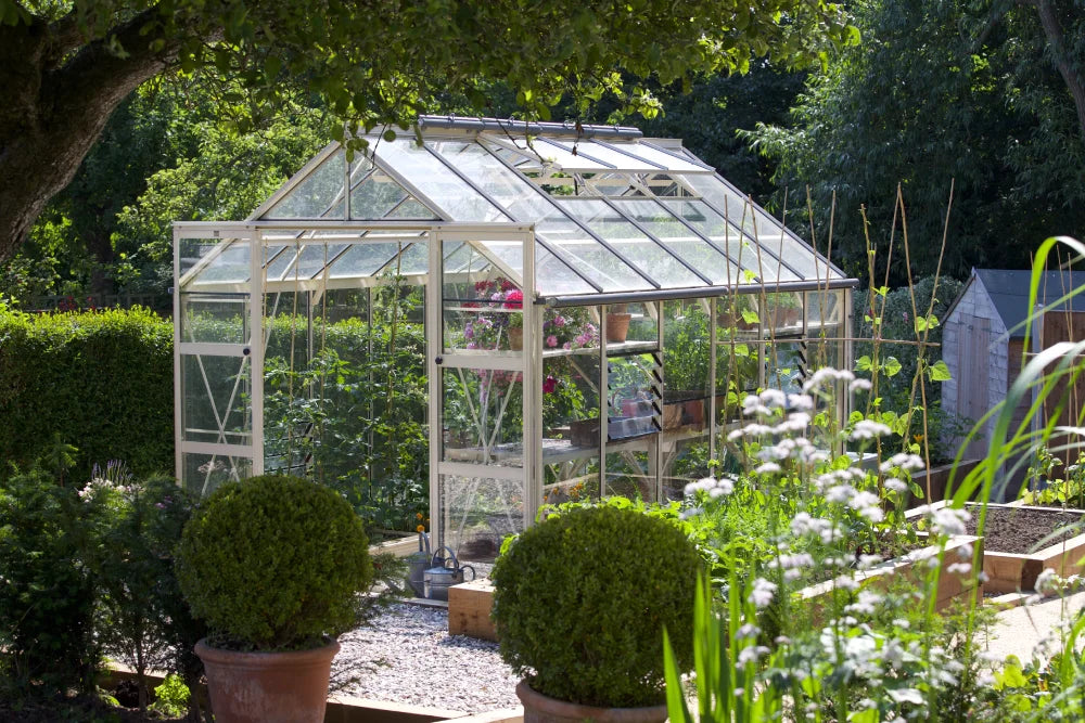 A glass Rhino Greenhouse filled with flourishing plants is centered in a lush garden, surrounded by trees and hedges, with potted shrubs and raised vegetable beds nearby.