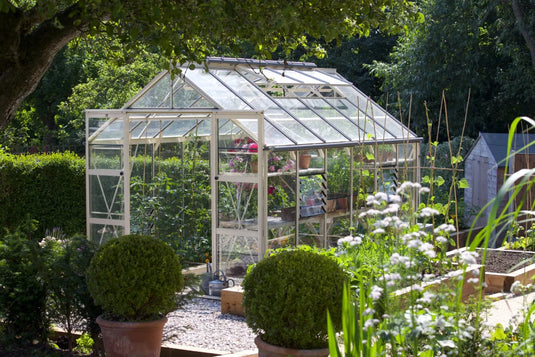 A glass Rhino Greenhouse filled with flourishing plants is centered in a lush garden, surrounded by trees and hedges, with potted shrubs and raised vegetable beds nearby.