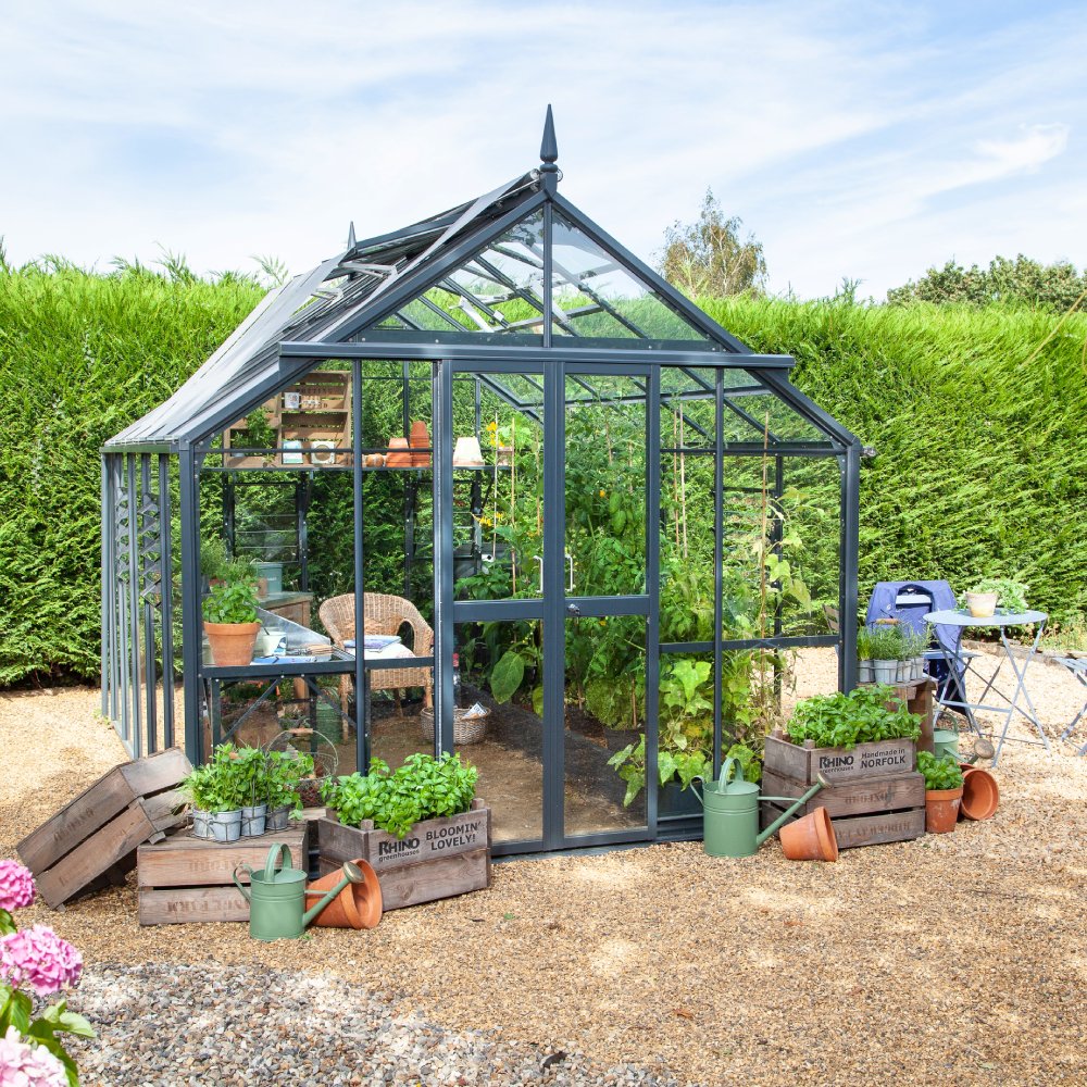 A glass Rhino Greenhouse houses lush plants and a wicker chair, surrounded by hedges. Wooden crates with Rhino, Norfolk, Bloomin’ Lovely hold potted plants, watering cans nearby on gravel.
