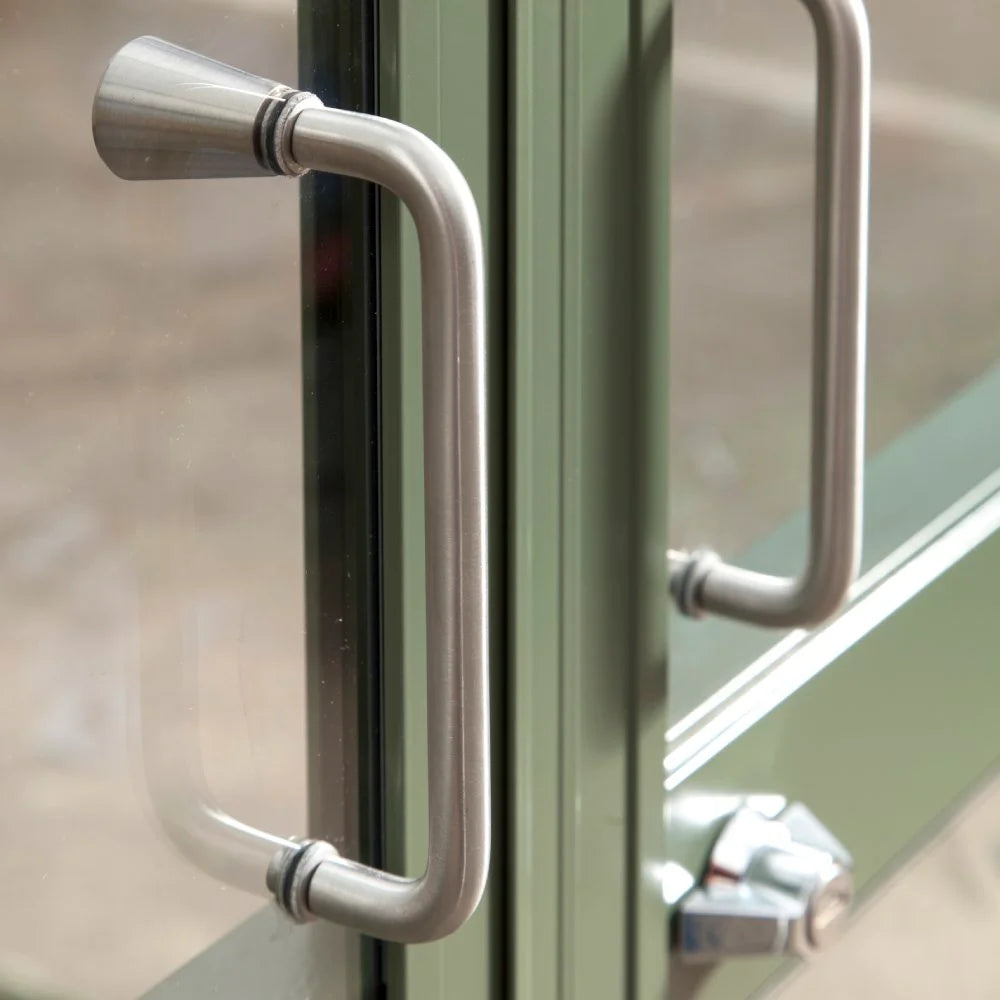 Metal handle affixed to green-framed glass door, slightly ajar, in a Rhino Greenhouse. The handle is cylindrical and brushed, with a lock visible below in a garden setting.