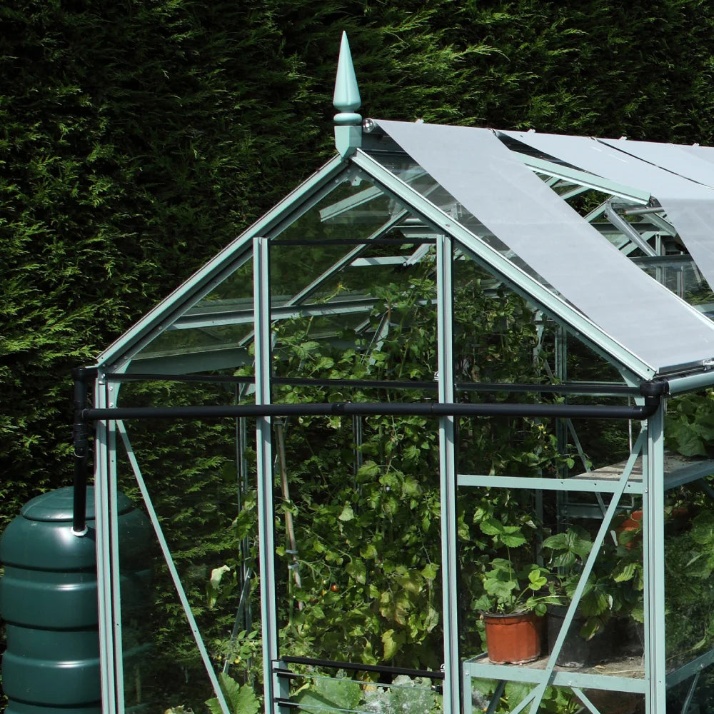 A glass Rhino Greenhouse with triangular roof panels houses various plants in pots, surrounded by lush greenery. A water barrel and a fence with a gate are visible nearby.