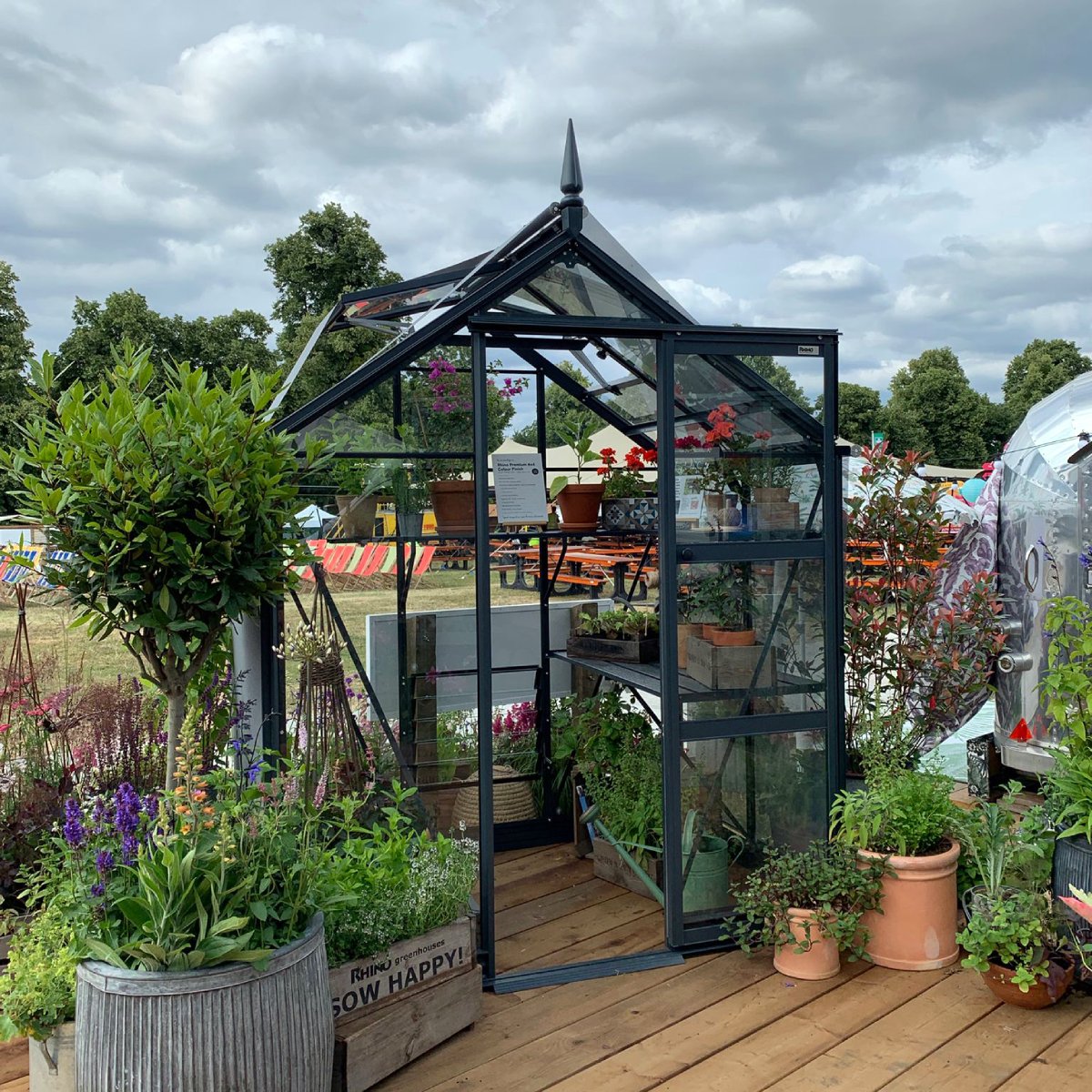 A glass Rhino Greenhouse with an open door displays potted plants and flowers inside, surrounded by more plants in various containers. It's set on a wooden deck in a park-like setting.