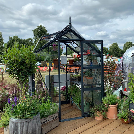 A glass Rhino Greenhouse with an open door displays potted plants and flowers inside, surrounded by more plants in various containers. It's set on a wooden deck in a park-like setting.