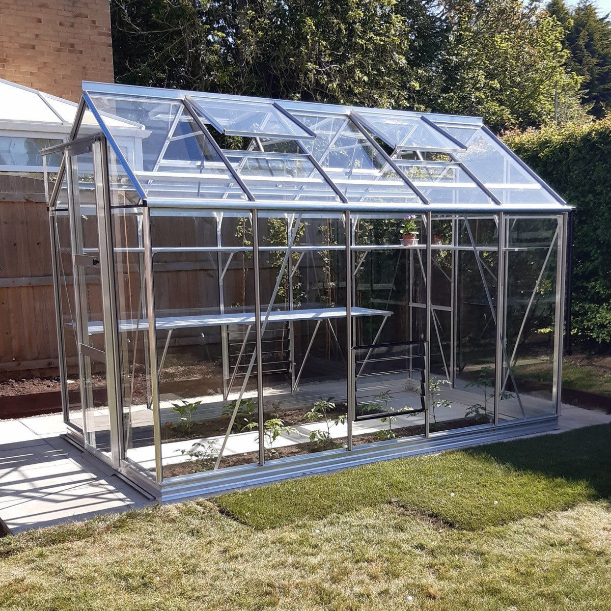 A glass Rhino Greenhouse with open windows sits on a paved area, surrounded by grass and trees, with some plants growing inside.