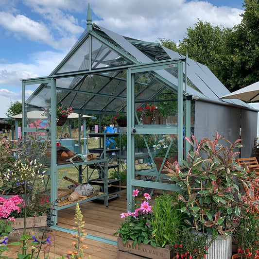 A Rhino Greenhouse with transparent panels and a peaked roof displays potted plants and gardening tools inside. Surrounded by colorful flowers and greenery on a wooden deck, under a partly cloudy sky.