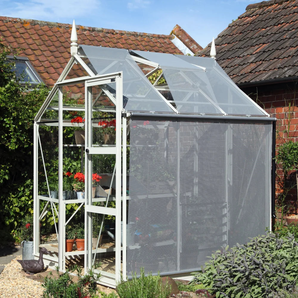 A white-framed Rhino Greenhouse with a mesh roof protects vibrant red flowers on shelves. It's situated in a garden area with brick buildings and green foliage in the background.