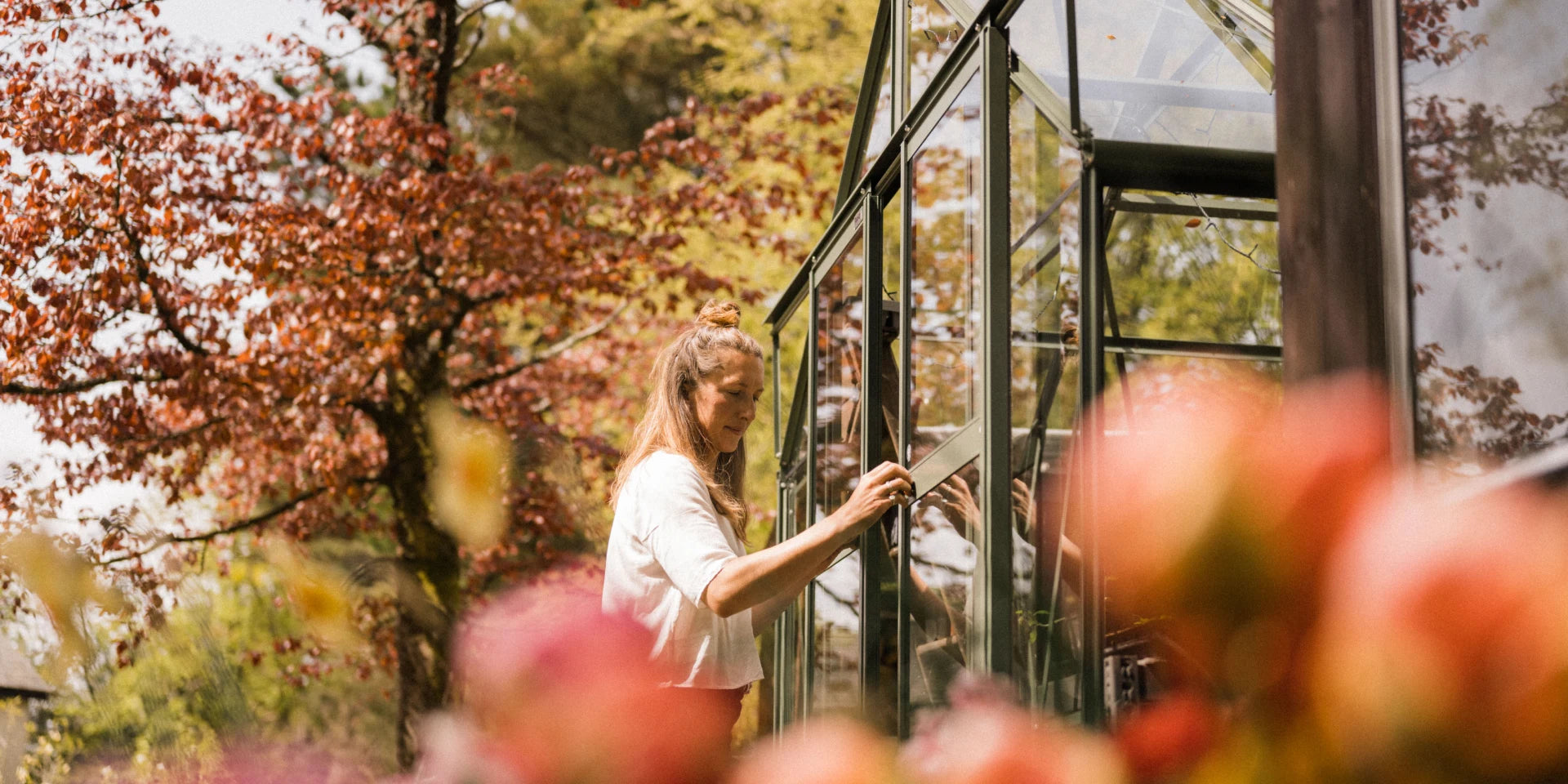 A person adjusts the window of a Rhino Greenhouse surrounded by vibrant autumn foliage and colorful flowers in a serene garden setting.