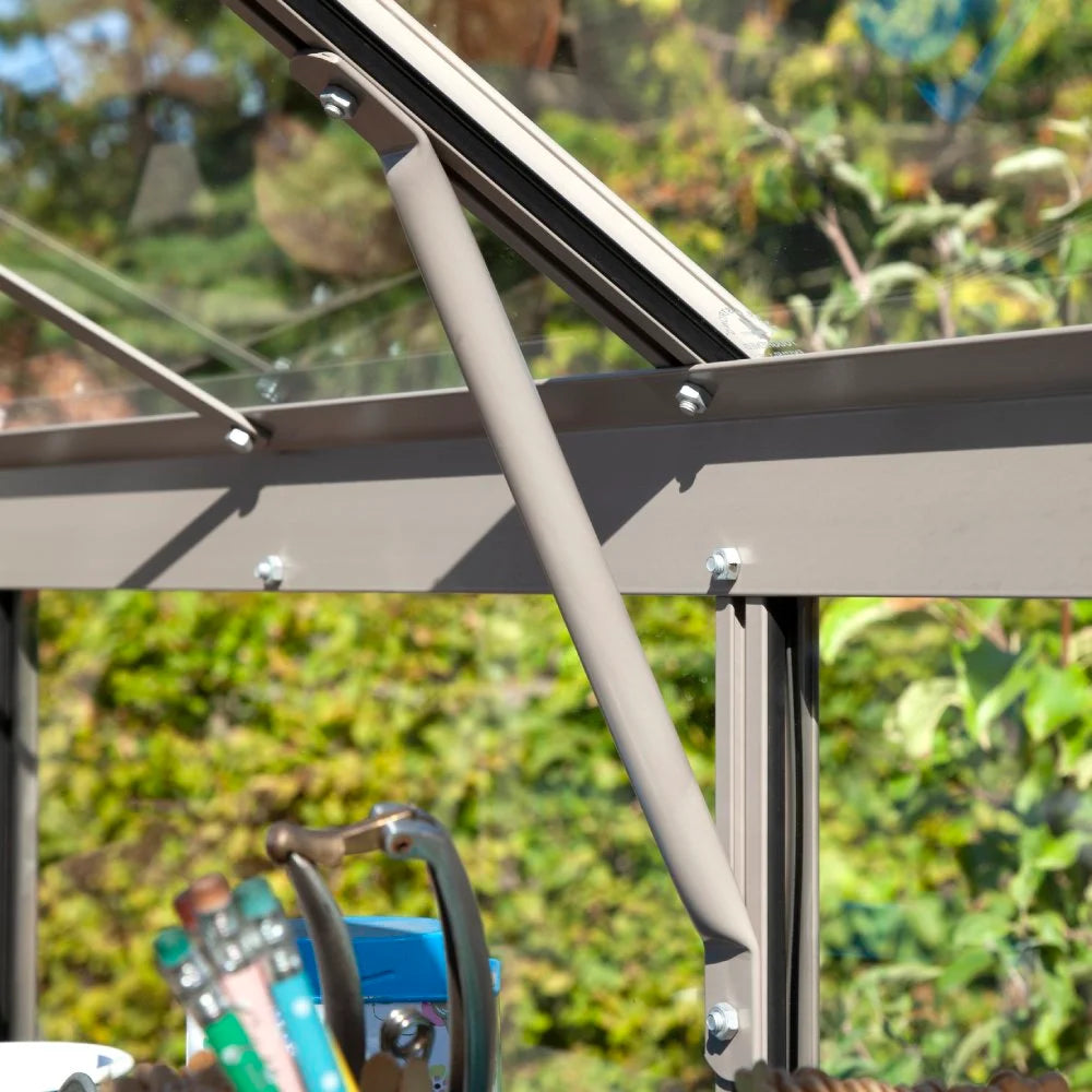 Metal support beam secures Rhino Greenhouse window, framed by bolts, in a lush garden setting. Gardening tools sit in the foreground, adding to the Rhino Greenhouse environment.